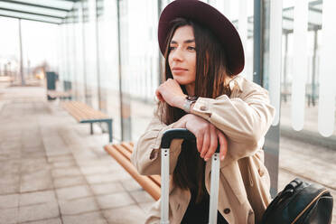 Nachdenkliches stilvolles Mädchen in Mantel und Hut sitzt an einer Haltestelle und wartet auf den Bus - CAVF83140