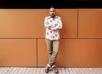 Bearded man leaning on wall with arms crossed against office building - CAVF83164