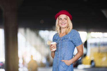 Porträt einer lächelnden blonden Frau mit Kaffee zum Mitnehmen in Jeanskleid und rotem Hut - DIGF12417