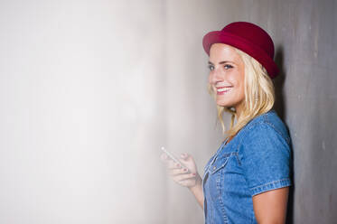 Porträt einer glücklichen jungen Frau mit Smartphone, die an der Wand lehnt - DIGF12420