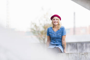 Portrait of happy young woman wearing denim dress sitting on a wall - DIGF12421