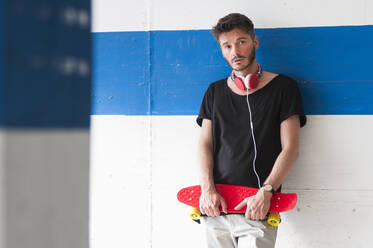 Portrait of man with skateboard leaning against wall - DIGF12423