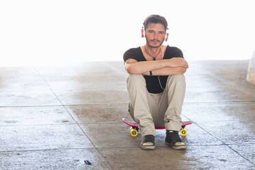 Portrait of man sitting on skateboard listening music with headphones - DIGF12424