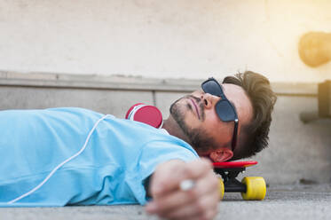 Portrait of man wearing sunglasses lying on the ground with head on his skateboard - DIGF12426