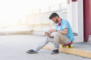 Porträt eines lächelnden Mannes mit Kopfhörern, der auf seinem Skateboard im Freien sitzt und ein digitales Tablet benutzt - DIGF12427