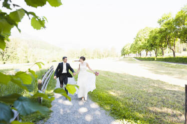 Cheerful newlywed couple holding hands while running on footpath against clear sky in park - DIGF12438