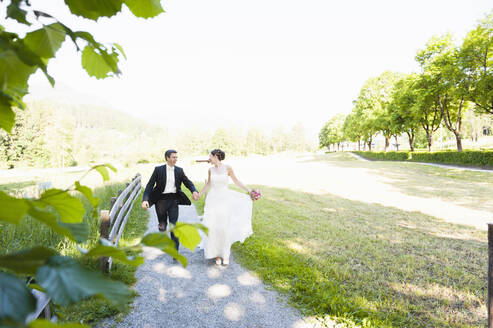 Fröhliches frisch verheiratetes Paar hält sich an den Händen, während es auf dem Fußweg gegen den klaren Himmel im Park läuft - DIGF12438