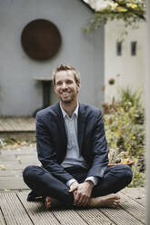 Laughing businessman sitting cross-legged in backyard - GUSF03822