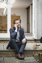 Businessman sitting in backyard, using smartphone while gardening - GUSF03825