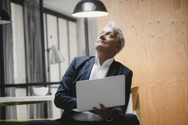 Älterer Geschäftsmann sitzt unter einer Deckenlampe, benutzt einen Laptop und schaut ins Licht - GUSF03826