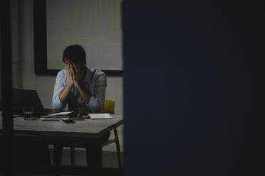 Erschöpfter Geschäftsmann, der sein Gesicht bedeckt, sitzt im Büro und arbeitet lange - GUSF03841