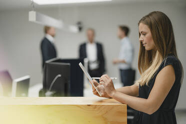 Geschäftsfrau in einem Büro stehend, auf einen Holztisch gestützt, mit einem digitalen Tablet - GUSF03878
