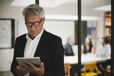 Senior businessman using digital tablet, coworkers working in background - GUSF03898