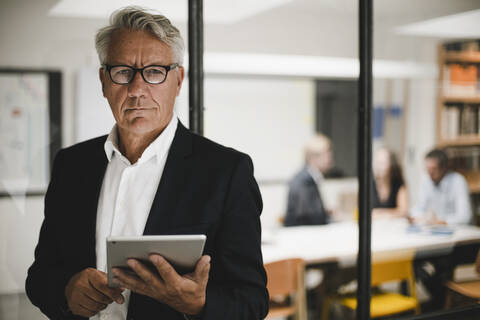 Älterer Geschäftsmann mit digitalem Tablet, Mitarbeiter arbeiten im Hintergrund, lizenzfreies Stockfoto