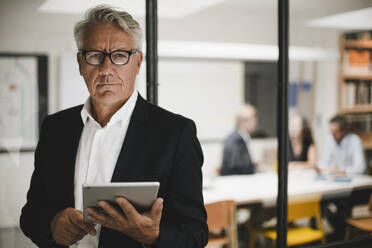 Senior businessman using digital tablet, coworkers working in background - GUSF03899