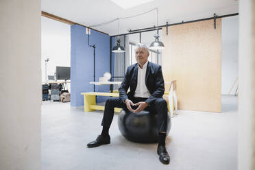 Senior businessman sitting on fitness ball, looking sceptical - GUSF03928