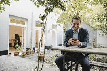 Smiling businessman sitting on bench in green backyard, using smartphone - GUSF03931