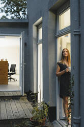 Geschäftsfrau steht in offener Bürotür und hält eine Tasse Kaffee - GUSF03954