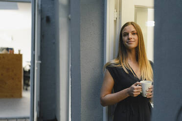 Businesswoman standing in open office door, holding cup of coffee - GUSF03955