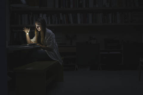 Junge Frau sitzt im Büro und liest nachts mit einer Taschenlampe in einem Buch, lizenzfreies Stockfoto