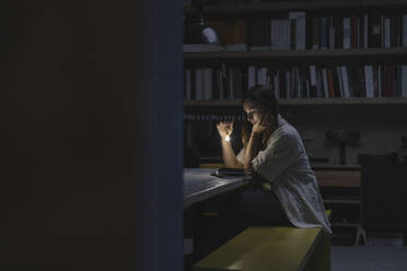 Young woman sitting in office, reading book at night with a torch - GUSF03963