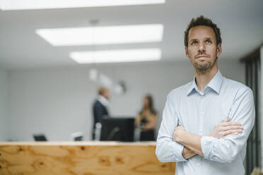 Businessman standing in open office door, with arms crossed , people working in background - GUSF04000