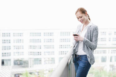 Porträt einer lächelnden jungen Frau auf einer Dachterrasse, die ein Smartphone benutzt - DIGF12455