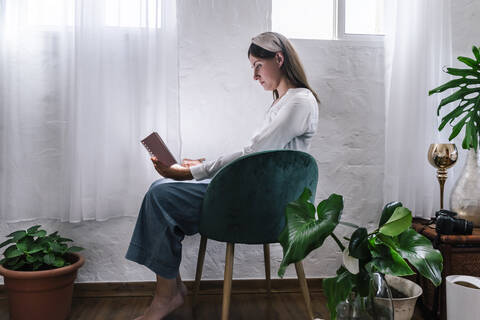 Relaxed woman reading book while sitting on chair by plants at home stock photo