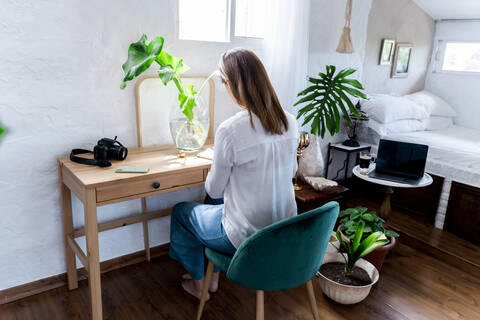 Frau mit langen braunen Haaren sitzt auf einem Stuhl am Schreibtisch im Schlafzimmer, lizenzfreies Stockfoto