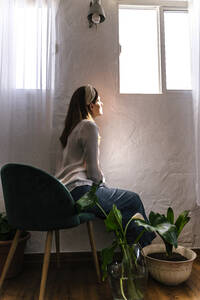 Frau sitzt auf einem Stuhl am Fenster und genießt das Sonnenlicht zu Hause - ERRF03899