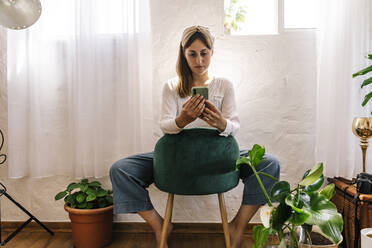 Beautiful woman using smart phone on chair at home - ERRF03901