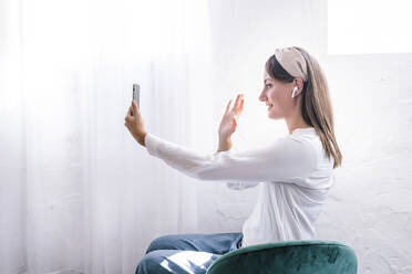 Lächelnde Frau winkt, während sie einen Videoanruf über ihr Smartphone zu Hause tätigt - ERRF03908