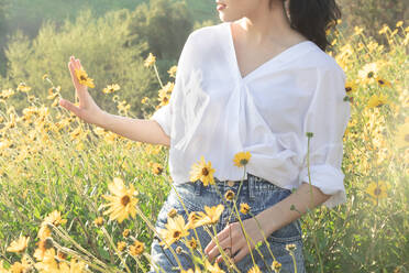 Brunette woman holding yellow daisy in flower field in the sunlight - CAVF83247