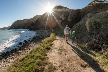 Vater und kleines Kind halten sich an einem sonnigen Tag auf einem Pfad in der Nähe des Meeres an den Händen - CAVF83300