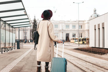 Stilvolles Mädchen mit einem Koffer steht an einer Haltestelle und wartet auf eine Straßenbahn - CAVF83326