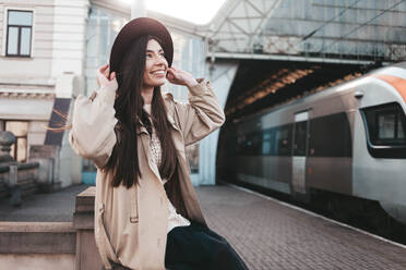 Lächelndes Mädchen mit Hut am Bahnhof, das auf die Abfahrt des Zuges wartet - CAVF83333