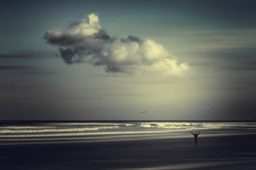 Frankreich, Nouvelle-Aquitaine, Contis, Wolken über der Silhouette eines Mannes, der mit erhobenen Armen am Strand von Contis Plage steht - DWIF01098