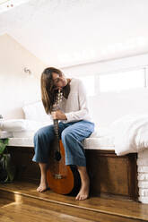 Woman with eyes closed sitting on bed with guitar relaxing - ERRF03923