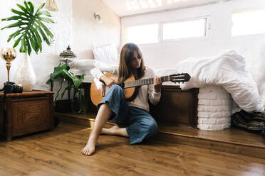 Woman sitting on the floor in front of bed playing guitar - ERRF03924