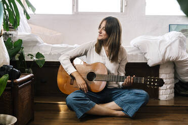 Woman sitting on the floor in front of bed with guitar - ERRF03926