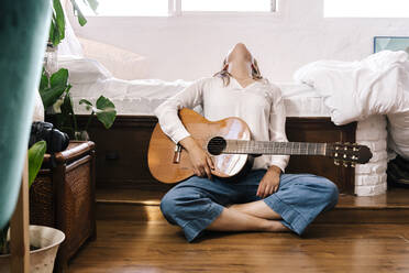 Frau mit Gitarre sitzt auf dem Boden vor dem Bett und lehnt sich zurück - ERRF03927