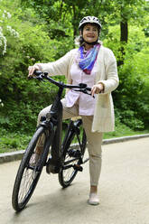 Thoughtful senior woman standing with electric bicycle on footpath in park - ECPF00931