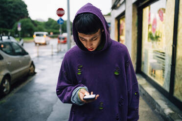 Junger Mann mit Kapuzenshirt, der auf der Straße stehend sein Smartphone benutzt - MEUF00609