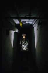 Man holding illuminated smart phone in dark corridor amidst old walls - MEUF00613