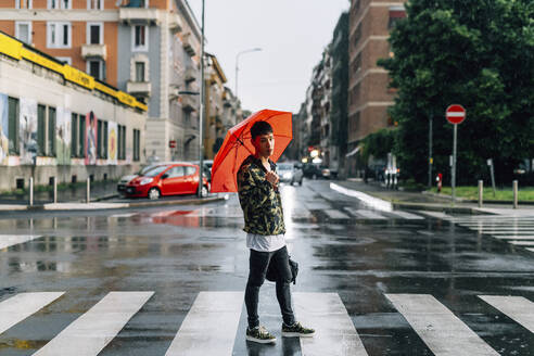 Junger Mann mit Regenschirm steht auf der Straße in der Stadt während der Regenzeit - MEUF00616