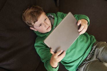 Junge benutzt digitales Tablet, während er zu Hause auf dem Sofa liegt - MMIF00273