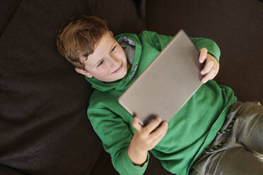Smiling boy using digital tablet while lying on sofa at home - MMIF00274