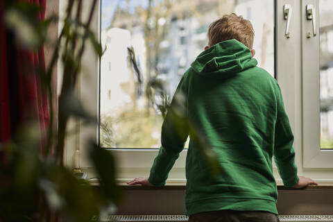 Junge in grünem Kapuzenshirt schaut zu Hause aus dem Fenster, lizenzfreies Stockfoto