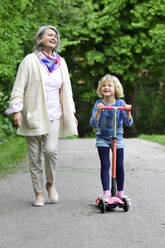 Happy senior woman walking with granddaughter riding push scooter in park - ECPF00941