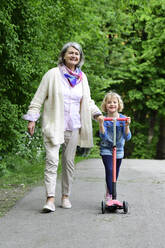 Smiling senior woman walking with granddaughter riding push scooter in park - ECPF00942